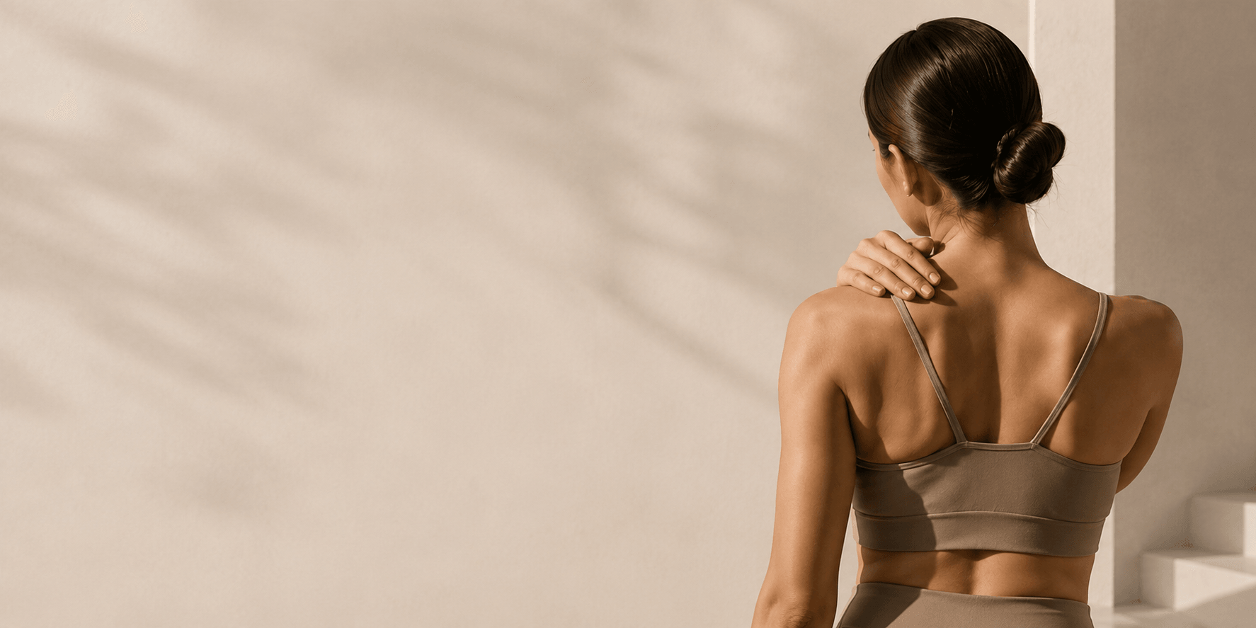 Woman in neutral activewear resting her hand on her shoulder, viewed from behind